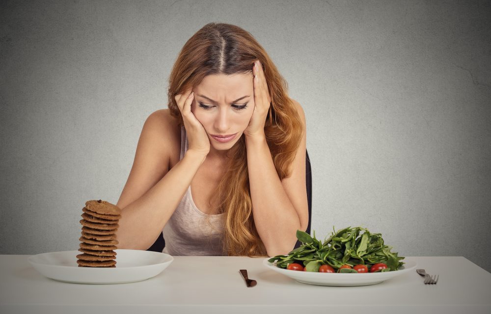 Young woman tired of diet restrictions deciding whether to eat healthy food or sweet cookies she is craving sitting at table isolated grey background. Human face expression emotion. Nutrition concept