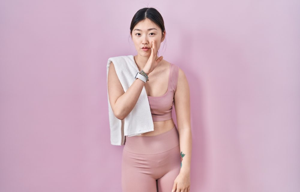 Chinese young woman wearing sportswear and towel hand on mouth telling secret rumor, whispering malicious talk conversation